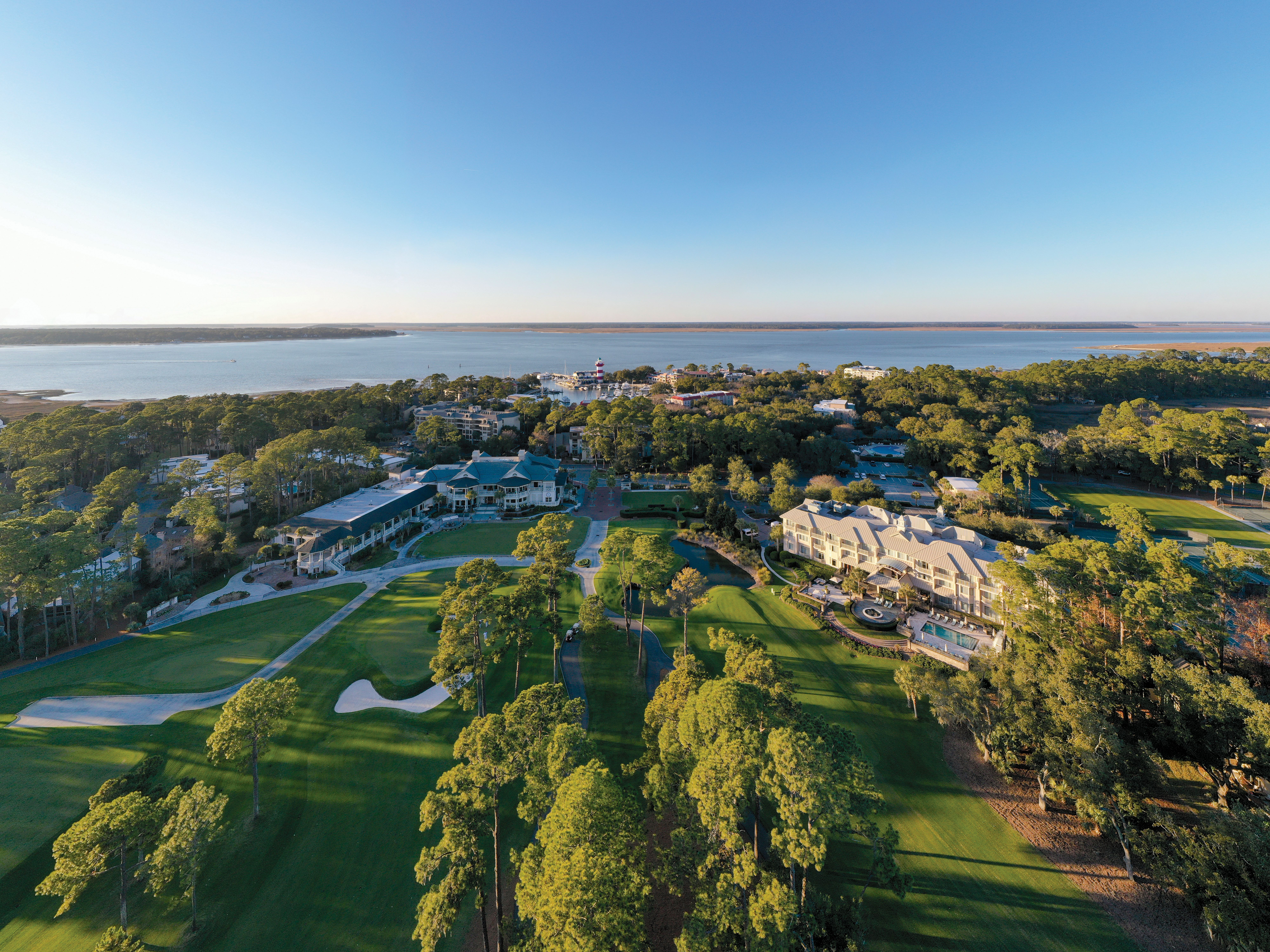 Aerial view of The Inn & Club at Harbour Town at The Sea Pines Resort on Hilton Head Island, overlooking Harbour Town Golf Links. Manicured fairways, pine trees, and resort accommodations are featured in the foreground, with Harbour Town Marina, the iconic red-and-white Harbour Town Lighthouse, and Calibogue Sound visible in the distance under a clear blue sky.