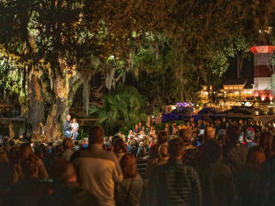 Guests gather beneath a sprawling live oak tree draped with Spanish moss for an evening holiday performance in Harbour Town. Warm lights illuminate the crowd and stage area, while the Harbour Town Lighthouse and marina glow in the background, creating a festive Lowcountry atmosphere.