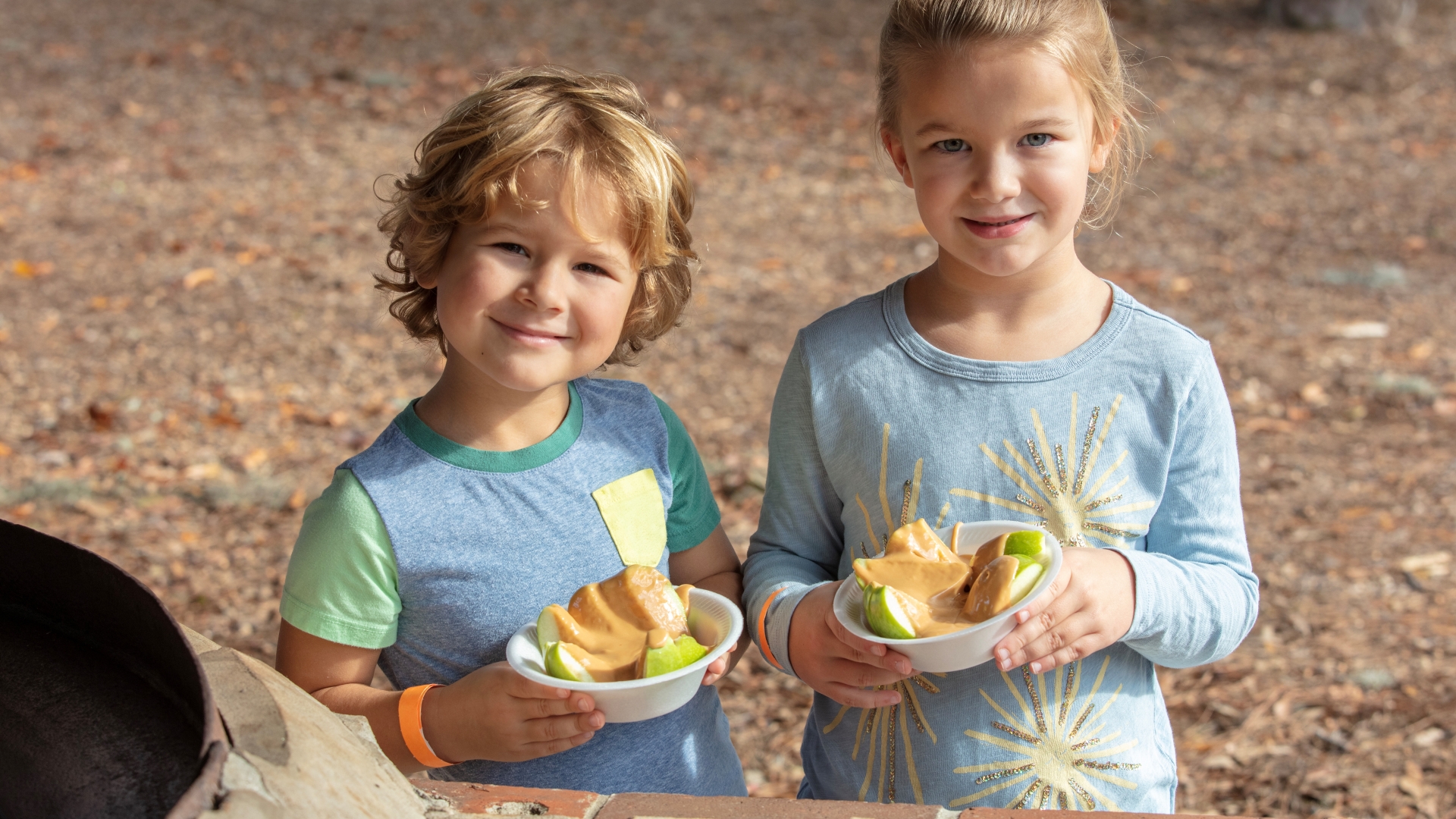 Two children hold bowls of apple slices and a caramel-colored dip while standing outdoors near a rustic stone counter. Surrounded by natural greenery and autumn foliage, the scene captures a family-friendly fall activity in a relaxed outdoor setting.