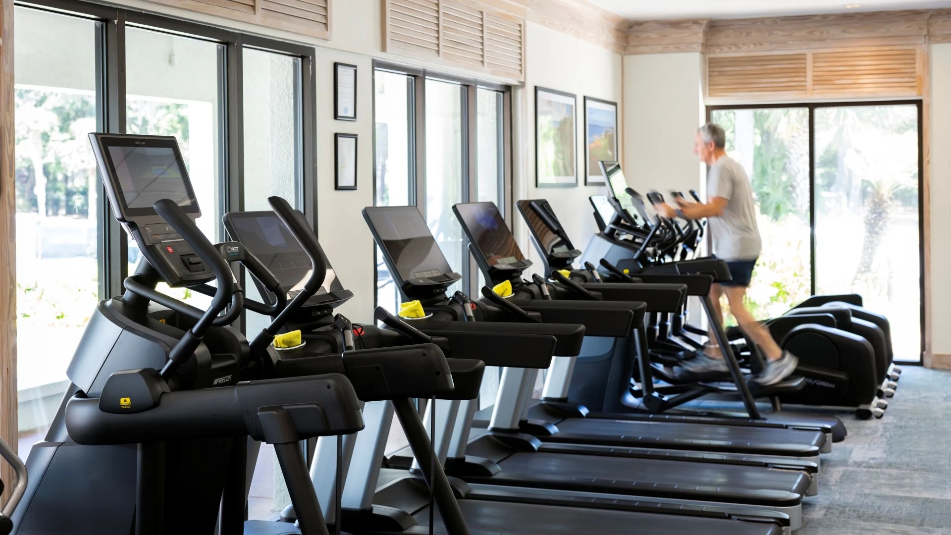 Interior of the fitness center at The Inn & Club at Harbour Town featuring modern treadmills, elliptical machines, and cardio equipment. Floor-to-ceiling windows provide natural light and views of the surrounding landscape while a guest exercises on a machine.