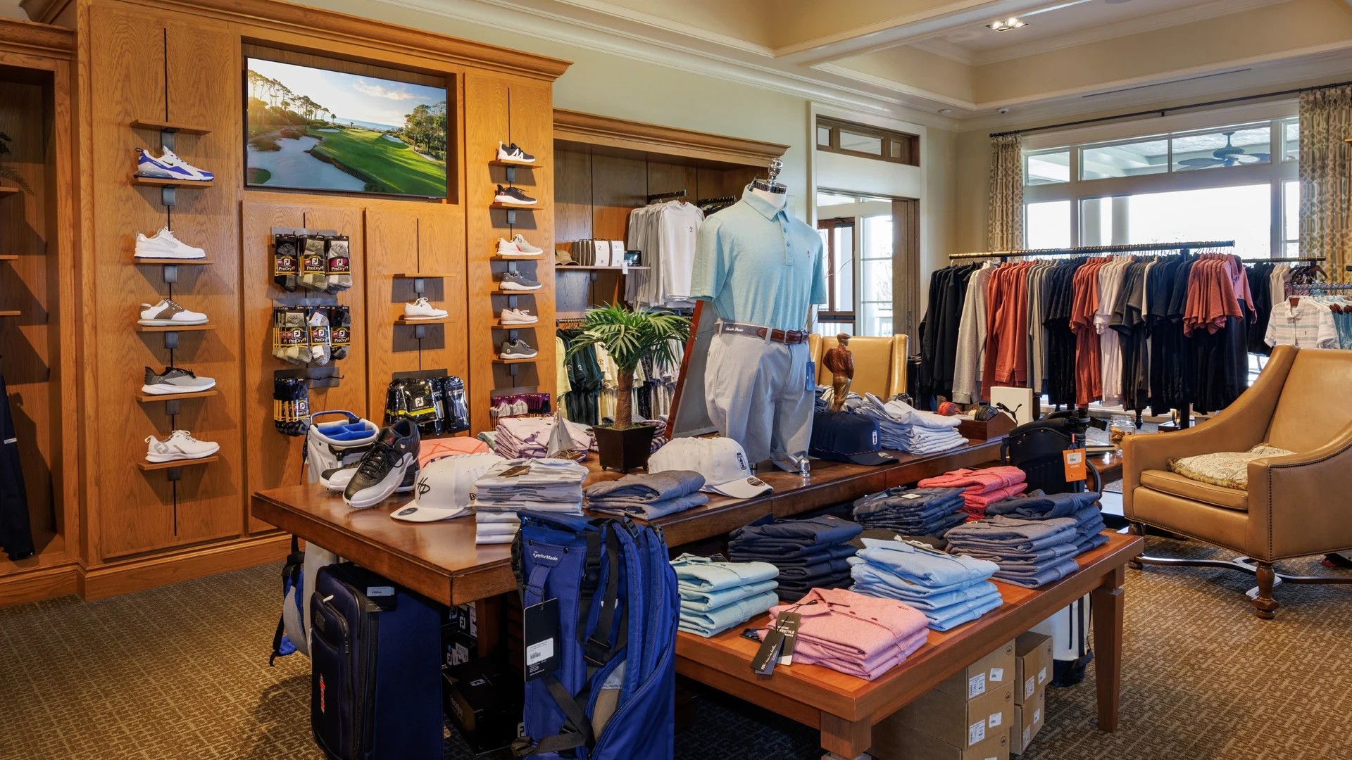 Interior of the Harbour Town Golf Links pro shop at The Sea Pines Resort, featuring displays of golf apparel, footwear, hats, luggage, and accessories. Folded shirts and merchandise are arranged on wooden tables, with clothing racks and golf gear showcased throughout the upscale retail space.