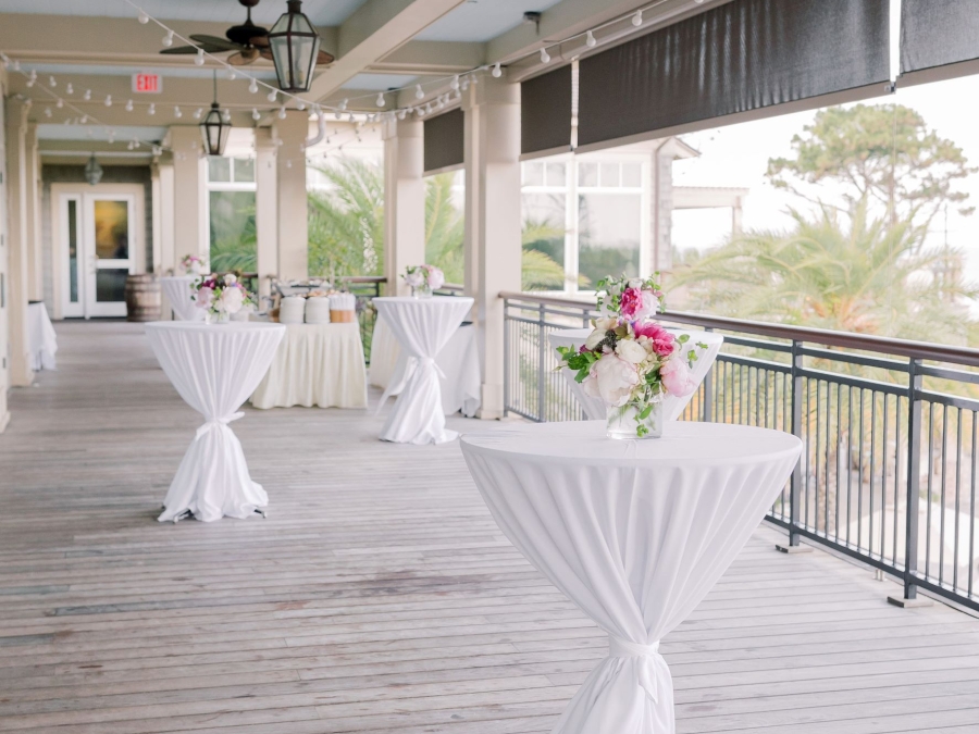 The deck outside of the Atlantic Room at the Sea Pines Beach Club set with high top round tables with floral arrangements for a wedding. 