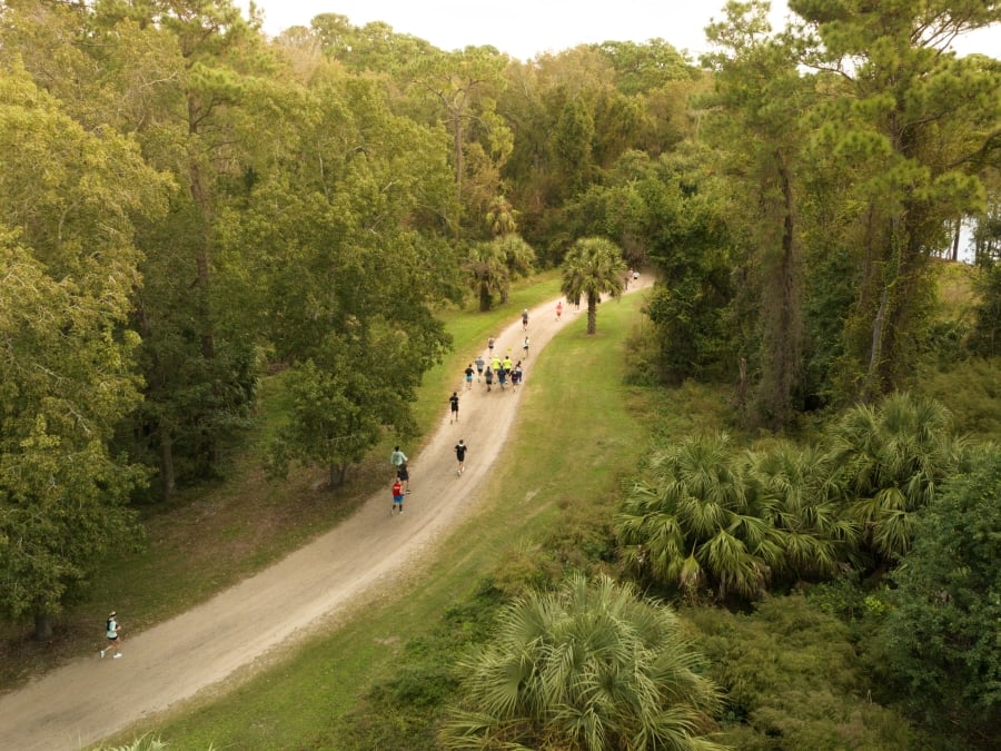 Aerial view of runners traveling along a winding unpaved trail through a dense maritime forest on Hilton Head Island. Tall pines, palmettos, and lush green vegetation line both sides of the path, creating a scenic natural corridor. Small groups of participants are spaced along the route as the trail curves through the landscape, highlighting an outdoor running event surrounded by the island’s distinctive Lowcountry environment.