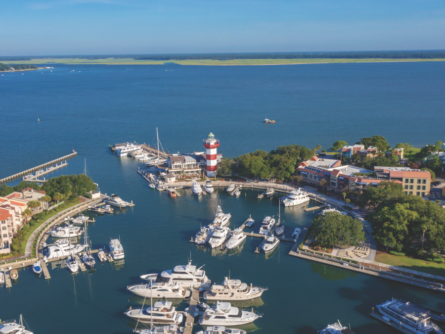Aerial view of Harbour Town Marina at The Sea Pines Resort on Hilton Head Island, featuring the iconic red-and-white Harbour Town Lighthouse, luxury yachts docked in the marina, waterfront shops and restaurants, and the calm waters of Calibogue Sound under a clear blue sky.