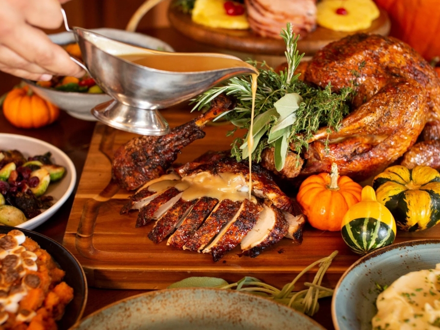 A Thanksgiving feast is displayed on a wooden table featuring roasted turkey, sliced turkey breast, seasonal side dishes, and festive fall décor. Gravy is poured over the carved turkey while mini pumpkins, fresh herbs, and colorful autumn dishes create a warm and inviting holiday dining scene.