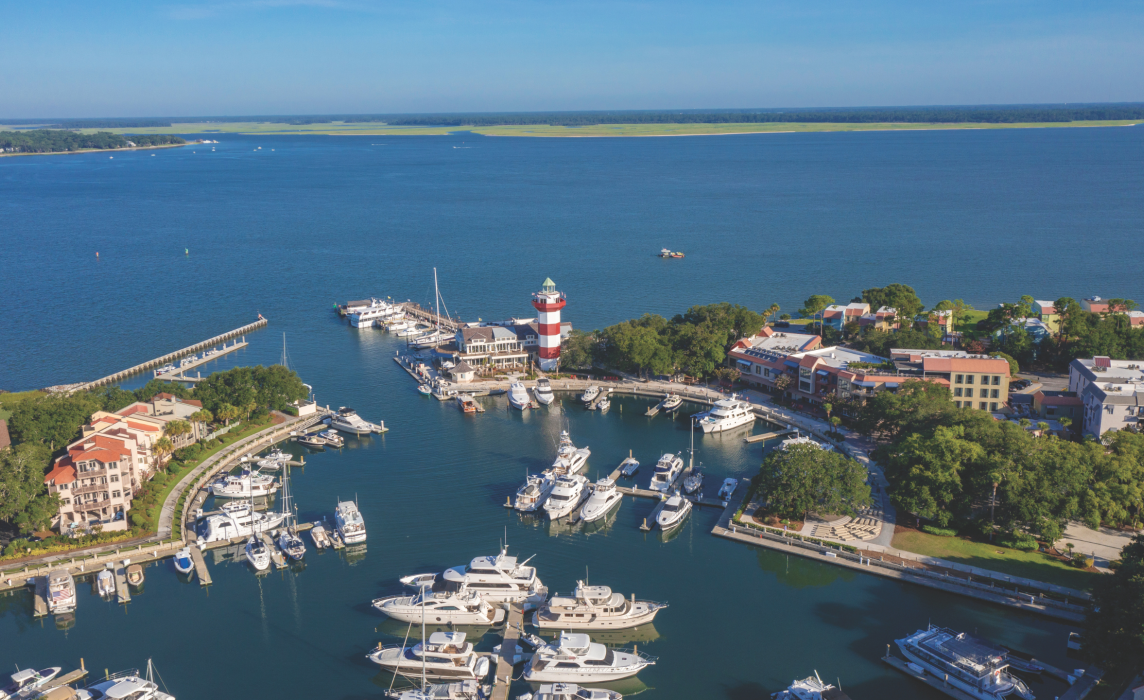 Aerial view of Harbour Town Marina at The Sea Pines Resort on Hilton Head Island, featuring the iconic red-and-white Harbour Town Lighthouse, luxury yachts docked in the marina, waterfront shops and restaurants, and the calm waters of Calibogue Sound under a clear blue sky.