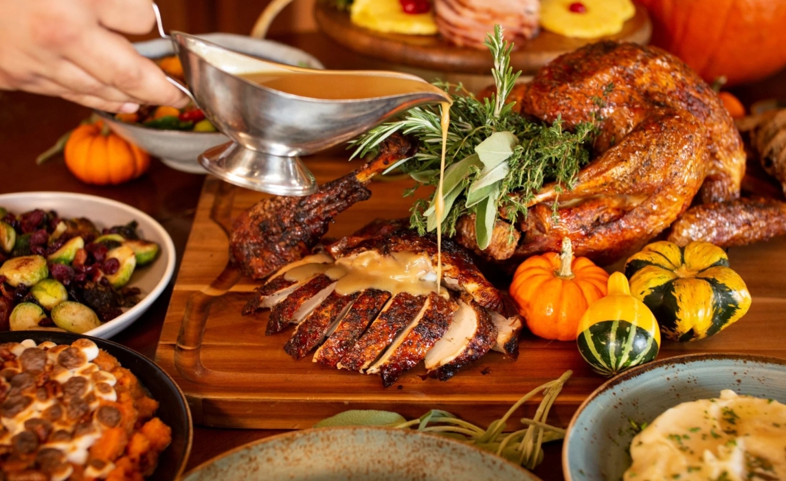 A Thanksgiving feast is displayed on a wooden table featuring roasted turkey, sliced turkey breast, seasonal side dishes, and festive fall décor. Gravy is poured over the carved turkey while mini pumpkins, fresh herbs, and colorful autumn dishes create a warm and inviting holiday dining scene.