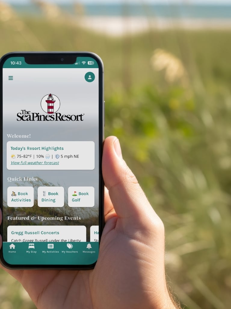 Hand holding a smartphone displaying The Sea Pines Resort mobile app, with quick links to book activities, dining, and golf. The device is positioned in front of a coastal dune landscape with beach grass and a blurred ocean view in the background.