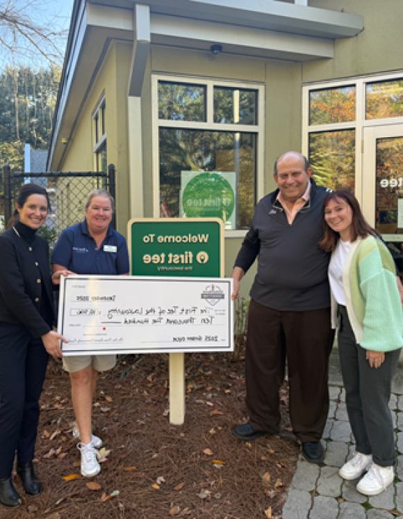 Four individuals stand outside a First Tee facility holding a ceremonial grant check awarded to First Tee as part of the 2025 grant cycle. A green and white First Tee sign is displayed near the entrance of the building, with landscaped grounds and a fenced area visible in the background. The presentation highlights community support and funding for youth development programs that teach life skills and character-building through golf.