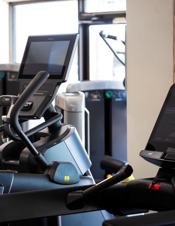Modern fitness center featuring cardio and strength-training equipment in a bright, sunlit space. An elliptical machine with a digital display is prominently positioned in the foreground, while additional fitness equipment, including treadmills and weight machines, fills the room.