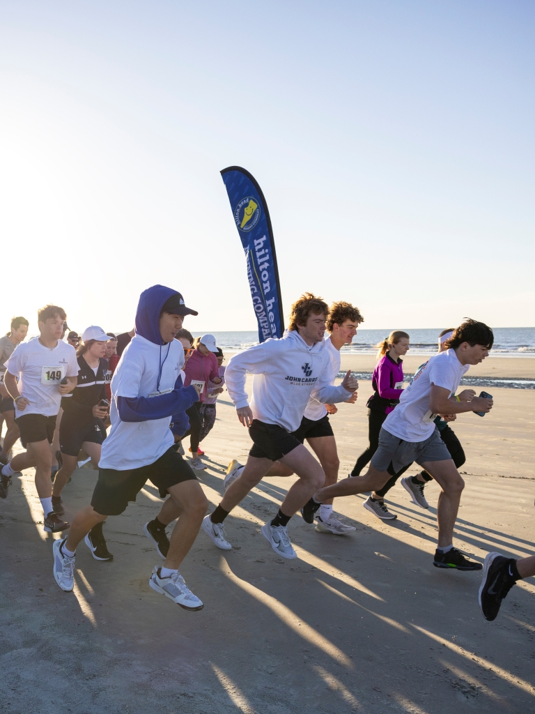 Participants take off from the starting line of a beach race as the sun rises over the Atlantic Ocean. Runners of all ages make their way across the sand beneath event banners, creating an energetic and community-focused atmosphere along Hilton Head Island’s shoreline.