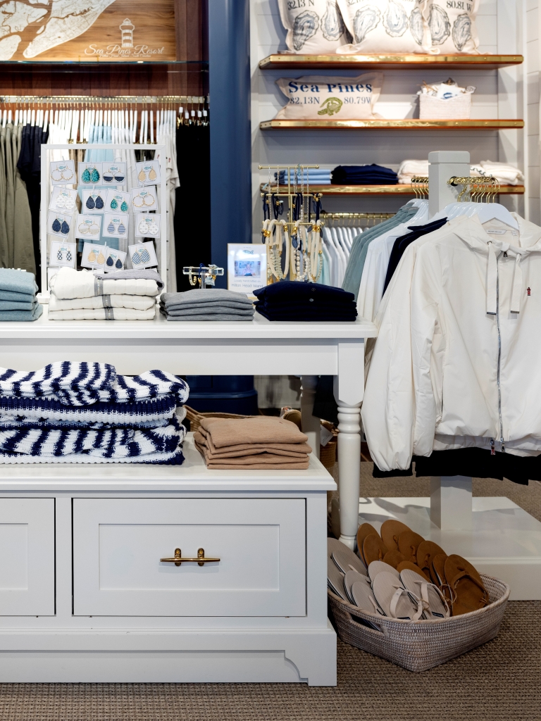 A boutique retail shop displays Sea Pines and Harbour Town apparel, accessories, and gifts arranged on shelves, racks, and display tables. Soft neutral colors, folded sweaters, jackets, hats, and coastal-inspired décor create an inviting shopping experience featuring resort-branded merchandise.